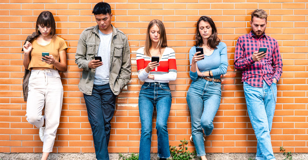 A diverse group of 20-somethings, standing against a wall, looking at their smartphones.