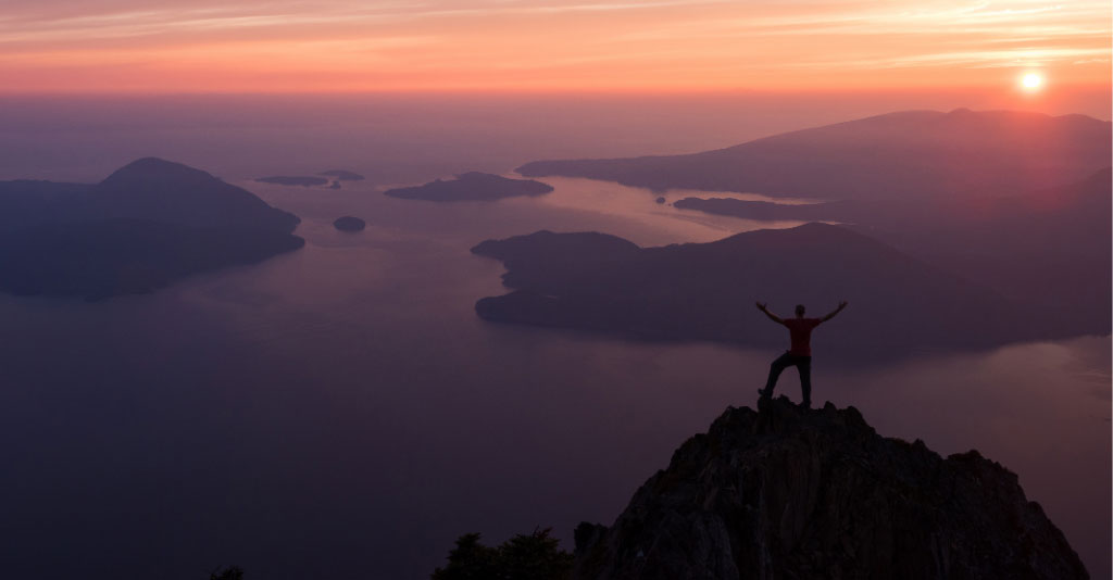 Silhouette of a person, holding their hands up in joy, on the top of a mountain overlooking the scene below at sunset.