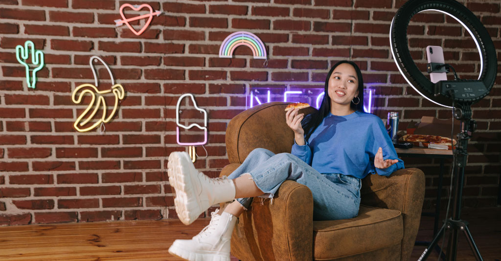 Young, female social media influencer recording herself on her phone with a brick background and neon signs.