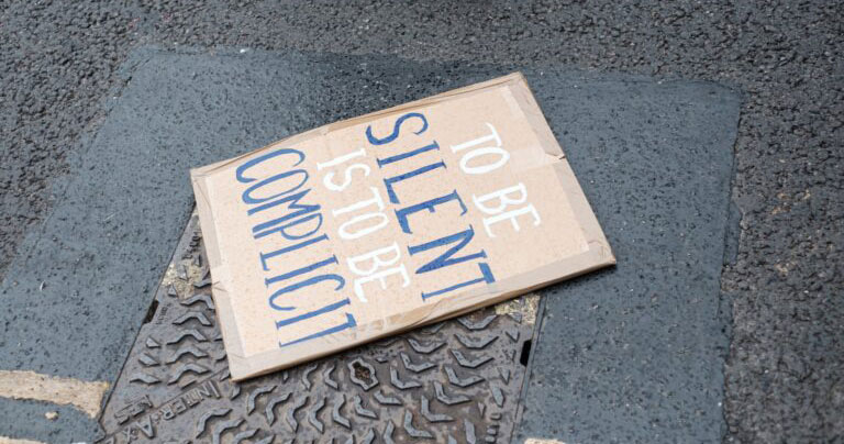 Cardboard sign left on the street that says "To Be Silent is To Be Complicit"